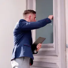 Mężczyzna w garniturze puka do drzwi, trzymając teczkę.